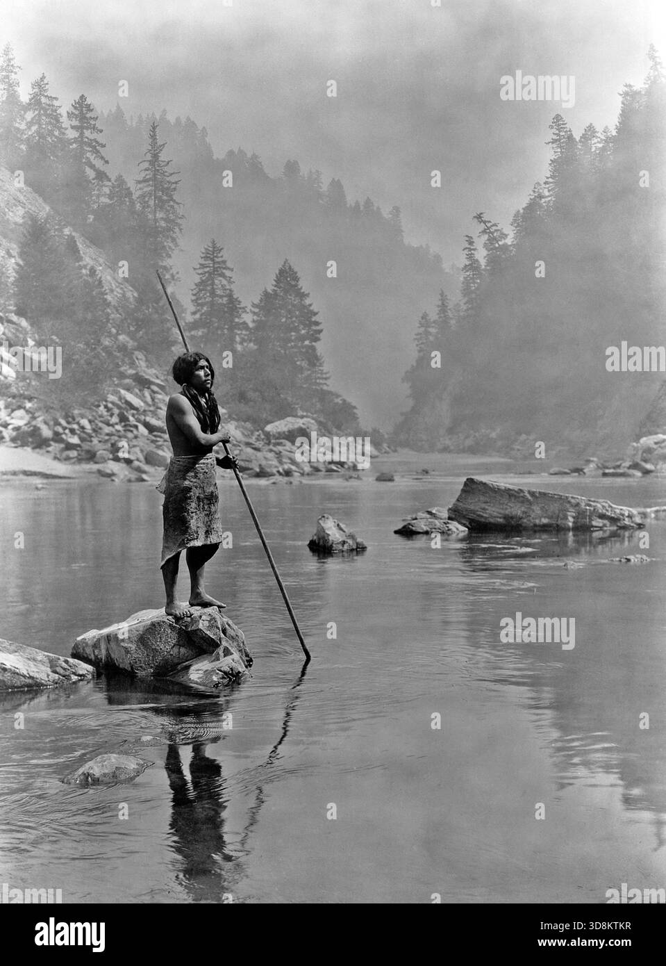 Une journée enfumée au Sugar Bowl--Hupa. Homme Hupa avec lance, debout sur la roche à mi-courant, en arrière-plan, le brouillard obscurcit partiellement les arbres sur les flancs des montagnes. Publié en 1924 dans le North American Indian / Banque D'Images