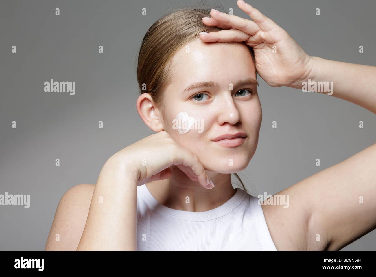 Jeune femme souriante avec une tache de crème hydratante sur les joues pose sur fond gris, illustrant des soins du visage doux quotidiens et une peau saine. Banque D'Images