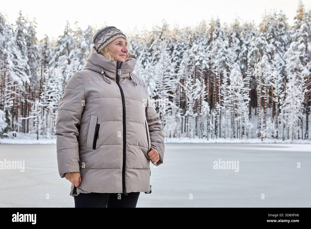 Femme se tient près d'un lac gelé, portant un manteau d'hiver et un chapeau tricoté, entourée d'arbres couverts de neige, profitant du paysage hivernal. Banque D'Images