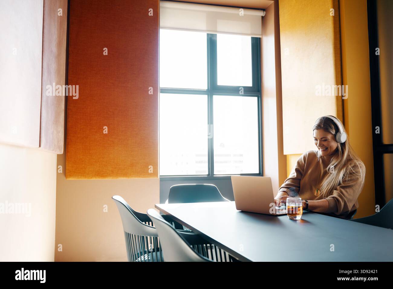 Une jeune femme est assise dans un bureau très éclairé, utilisant un ordinateur portable tout en portant des écouteurs. L'ambiance est professionnelle et accueillante, avec un accent sur la conce Banque D'Images