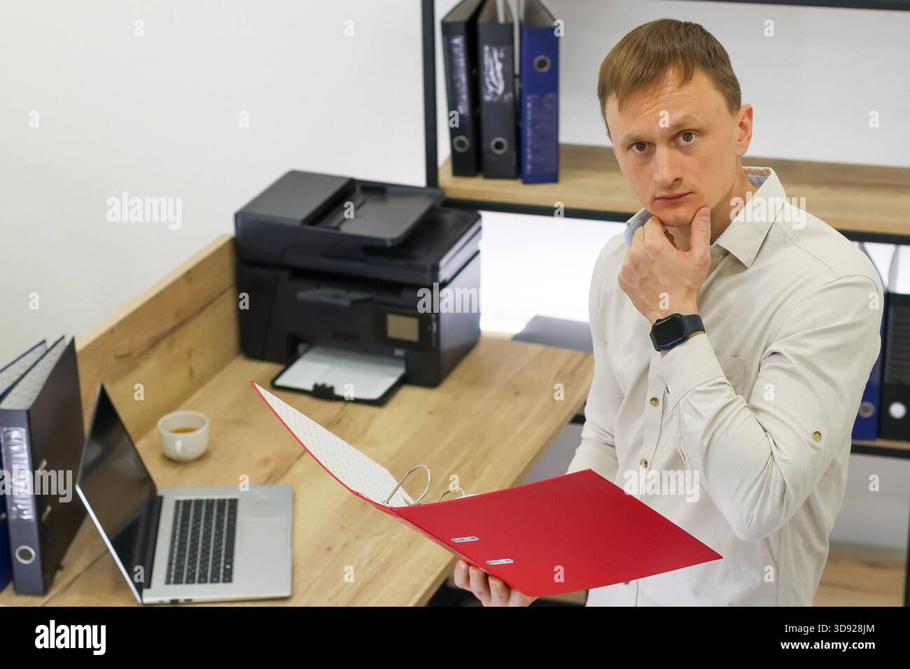 Homme en chemise beige assis à son bureau, la main sur le menton, examinant le contenu du dossier rouge au milieu des fournitures de bureau comme l'imprimante et l'ordinateur portable. Banque D'Images