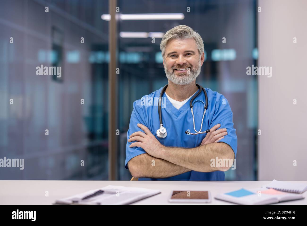 Professionnel de la santé portant des gommages bleus et stéthoscope, assis à un bureau avec les bras croisés, souriant à la caméra dans un hôpital ou une clinique moderne Banque D'Images