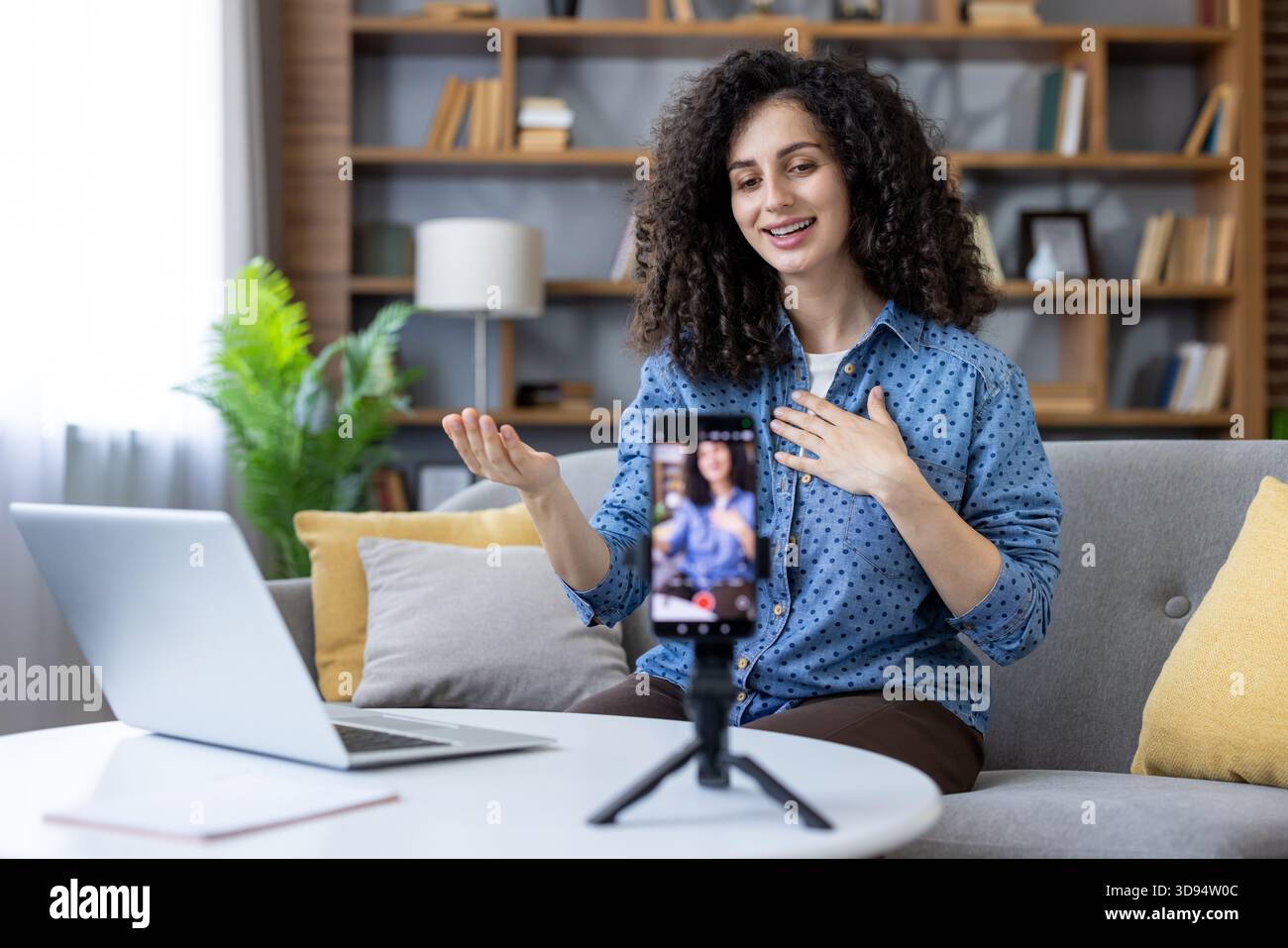 Femme créant du contenu en ligne, parlant à la caméra pendant un live ream réussi ou un appel vidéo de la maison, en utilisant un smartphone sur un trépied avec un ordinateur portable présent Banque D'Images