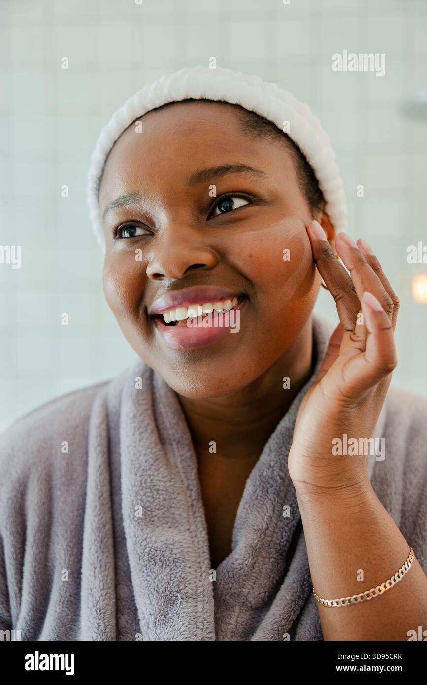 Femme afro-américaine lissant la crème dans la joue avec le bout des doigts dans la salle de bains portant un peignoir Banque D'Images