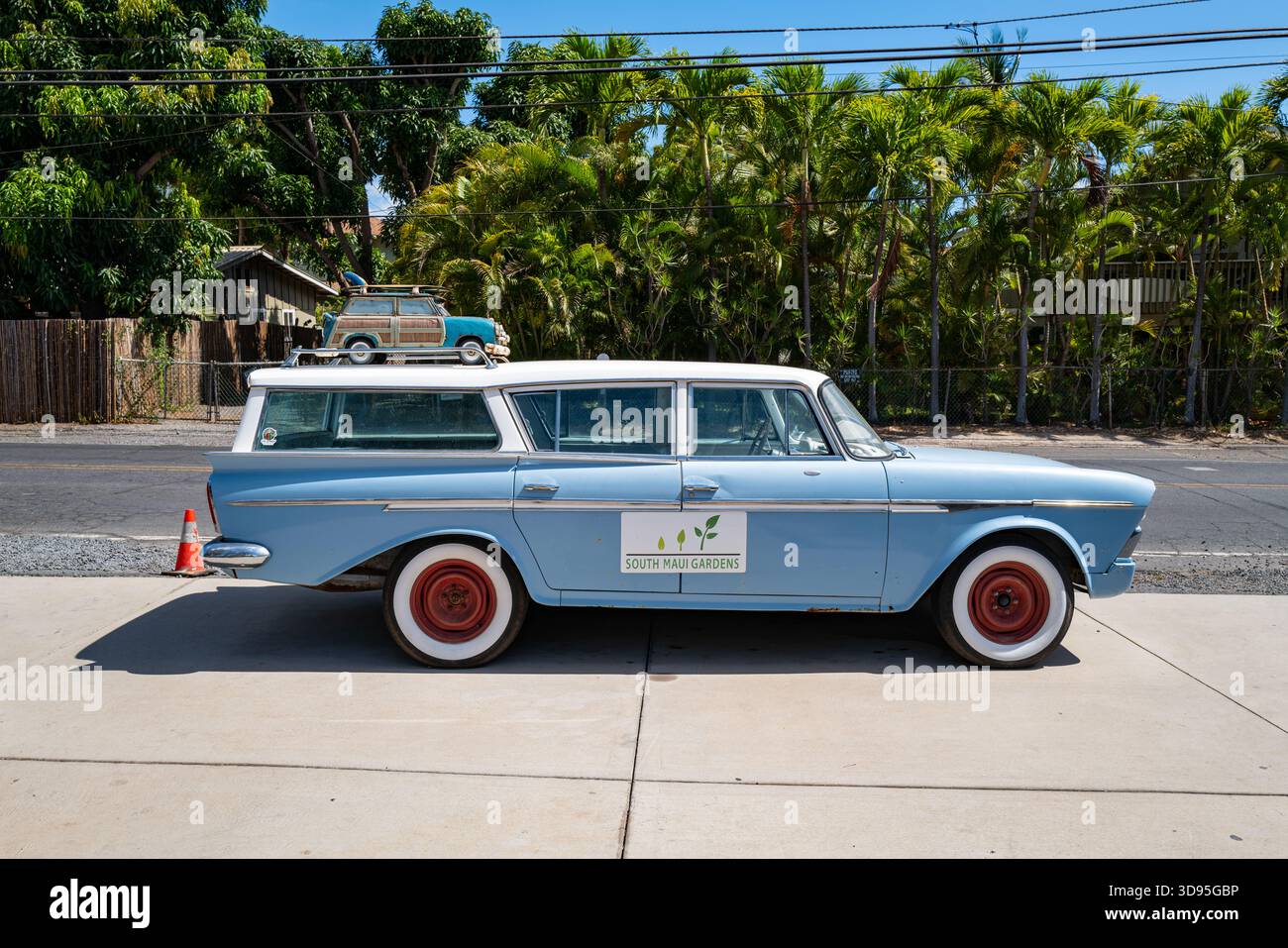 Wagon rétro bleu clair avec enjoliveurs rouges et panneau South Maui Gardens. Wagon de station boisé jouet avec planche de surf dans la rue par l'entrée. Banque D'Images