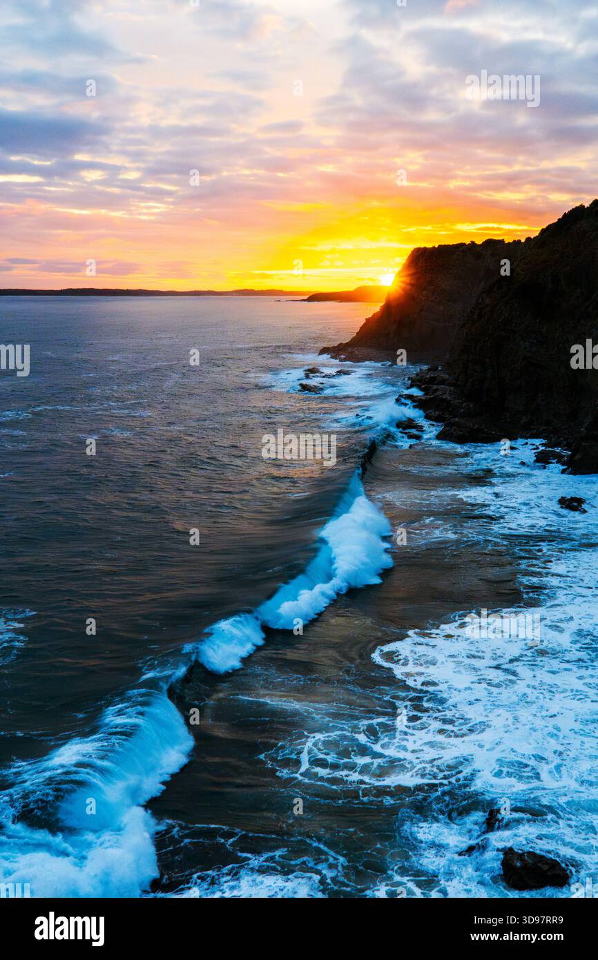 Les puissantes vagues bleues de l'océan s'écrasent contre une falaise sombre et accidentée lors d'un coucher de soleil coloré, illuminant le paysage côtier spectaculaire. Banque D'Images
