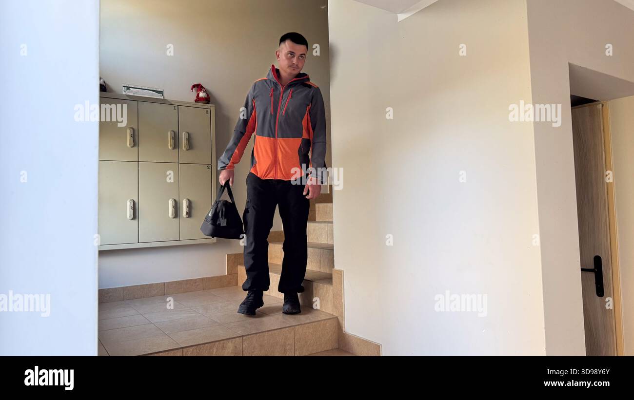 Un homme dans une veste grise et orange descend les escaliers carrelés dans un couloir d'appartement, transportant un sac de sport noir près d'un casier mural et Lig Banque D'Images