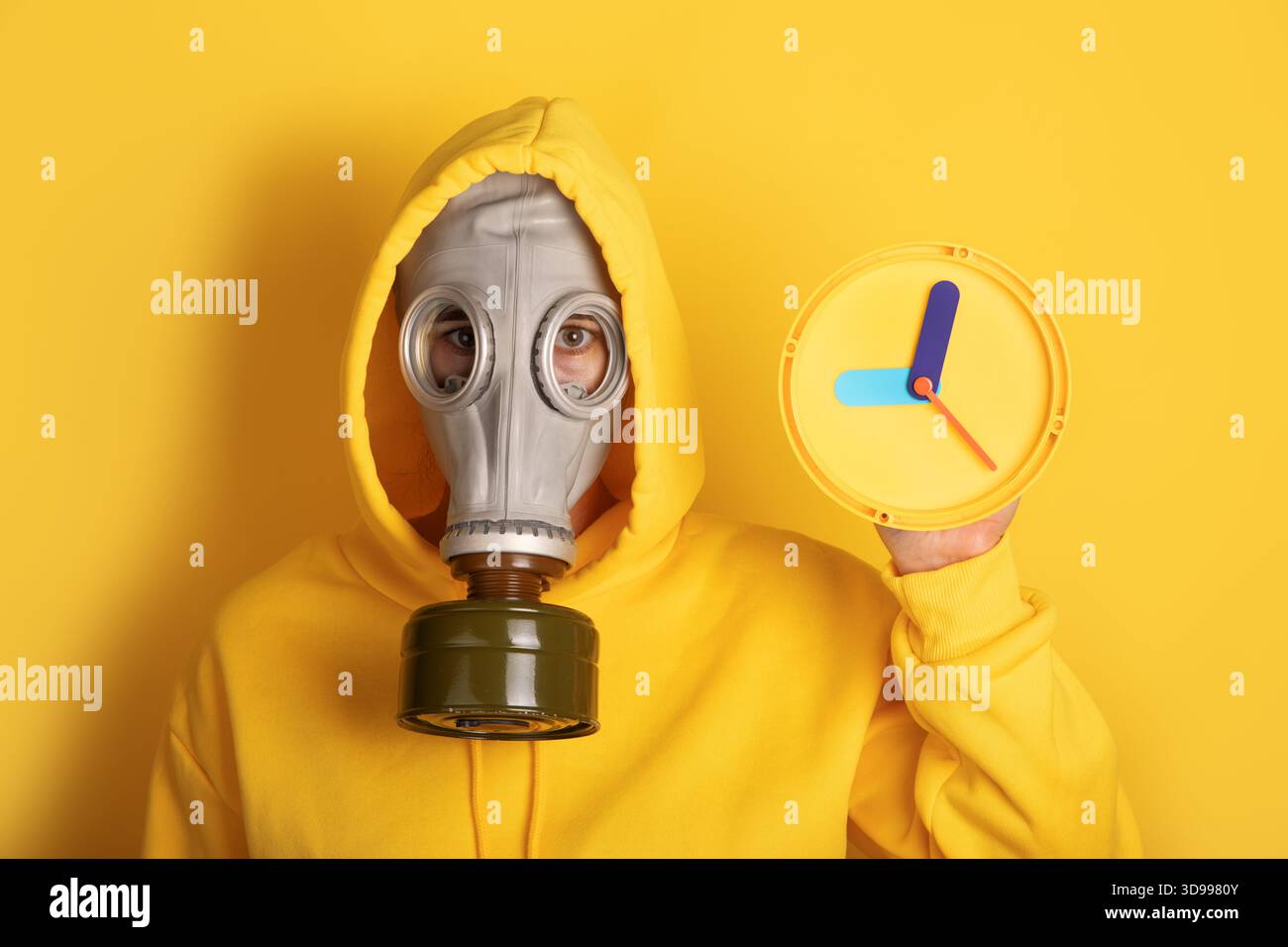 Personne en sweat à capuche jaune et masque à gaz tenant l'horloge colorée sur fond jaune vif, symbolisant l'urgence et la protection. Banque D'Images