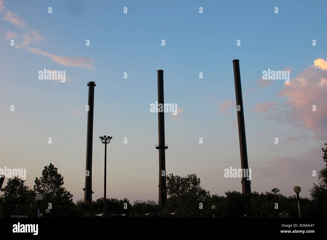 Une photographie de trois hautes cheminées d'usine s'élevant au-dessus du feuillage vert contre le ciel du soir. Nuages doux et teintes chaudes du soleil couchant ajoutent atm Banque D'Images