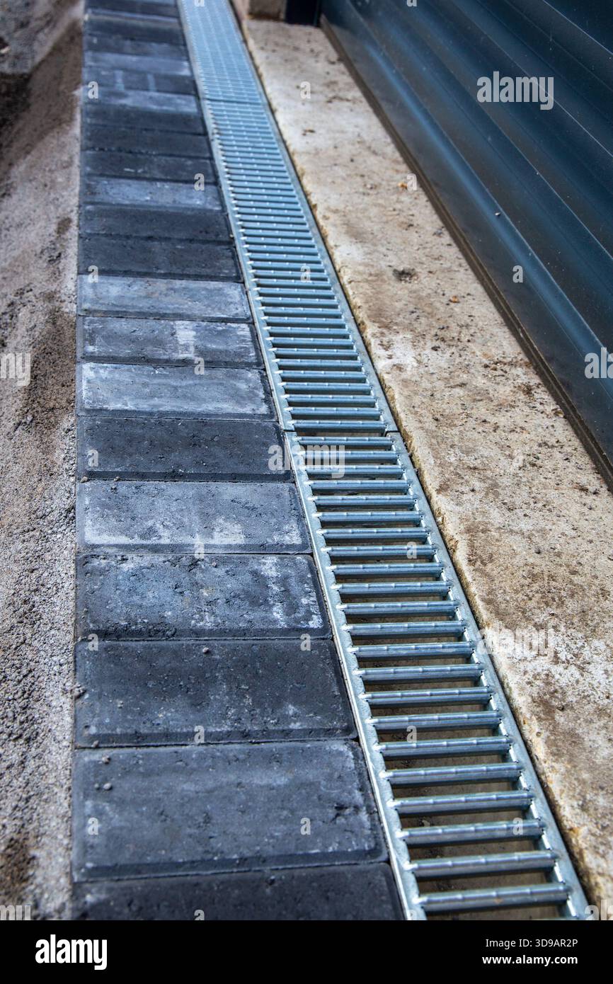 Vue rapprochée d'une grille de drainage métallique nouvellement installée à côté de pavés sombres et d'une surface en béton à l'extérieur d'une porte de garage. Banque D'Images