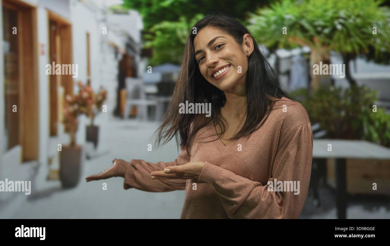 Femme souriant et présentant avec les mains sur le café de rue ensoleillé portant un pull rose col v et cheveux lâches ; confiance ludique. Banque D'Images