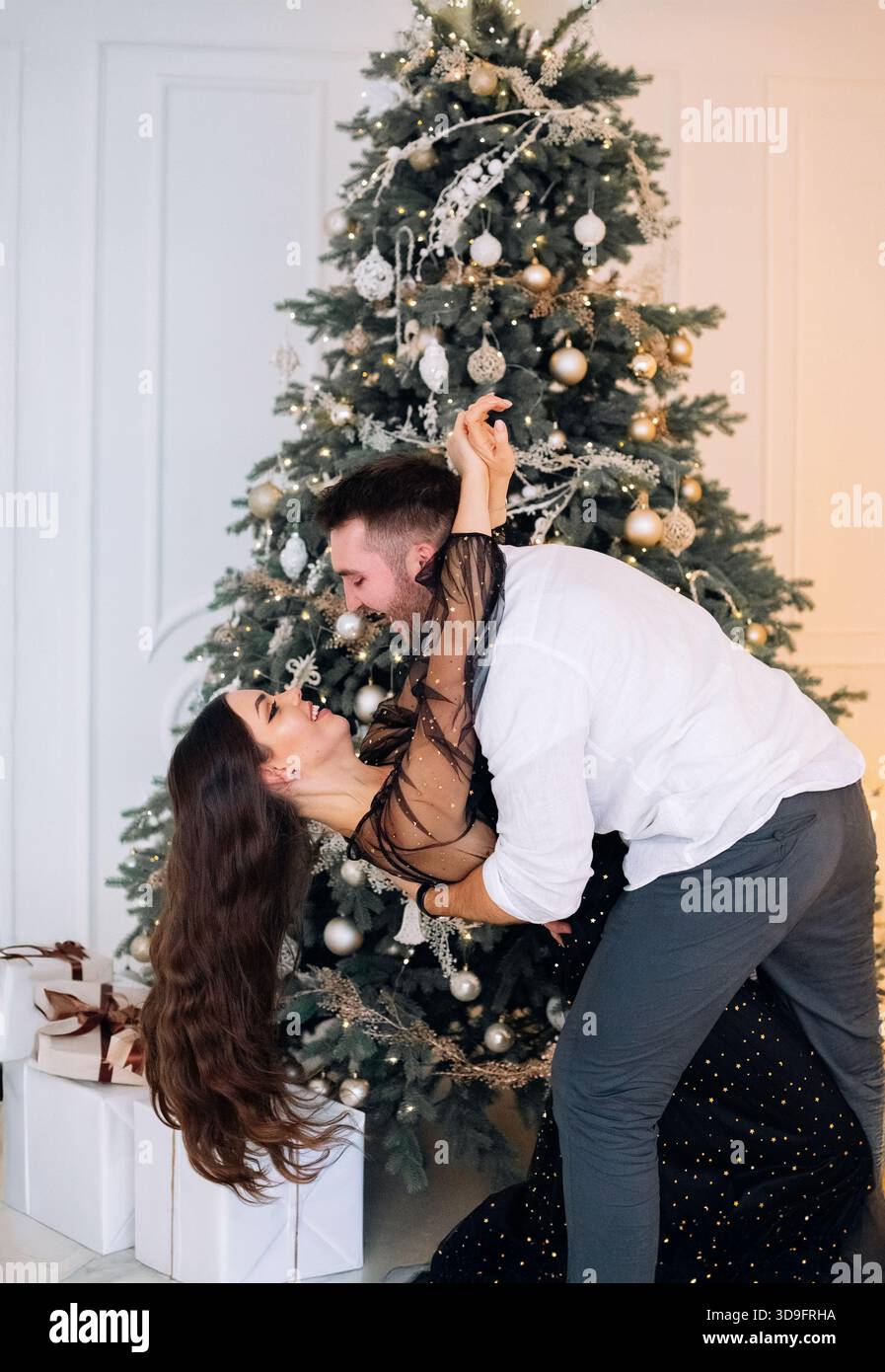 Une femme dans une élégante robe noire et son partenaire profitent d'une danse amusante près d'un sapin de Noël joliment décoré avec des lumières et des ornements, célébrant Banque D'Images