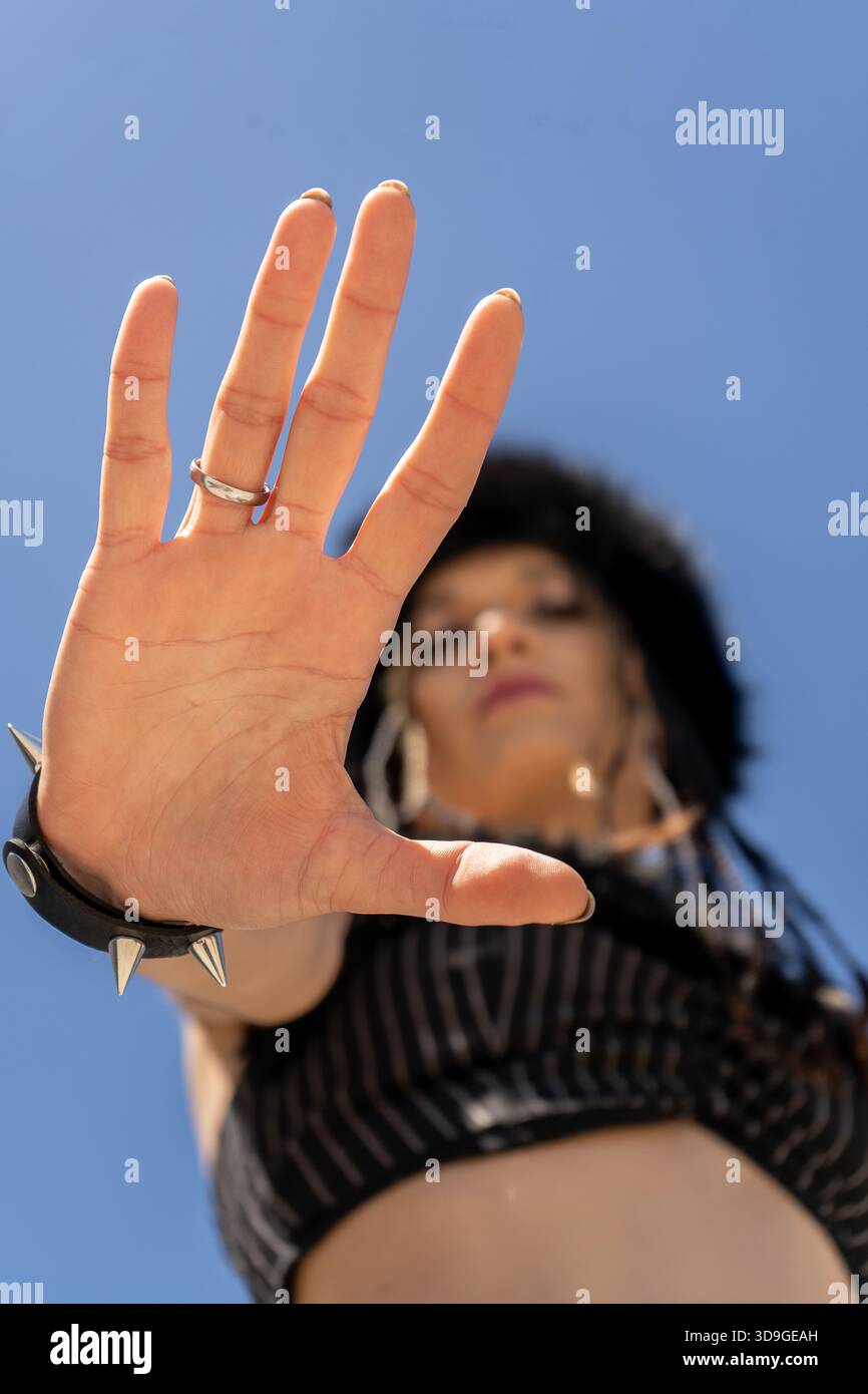 Femme projetant un puissant geste d'arrêt avec sa main, portant un bracelet à pointes et montrant la défiance contre un ciel bleu Banque D'Images