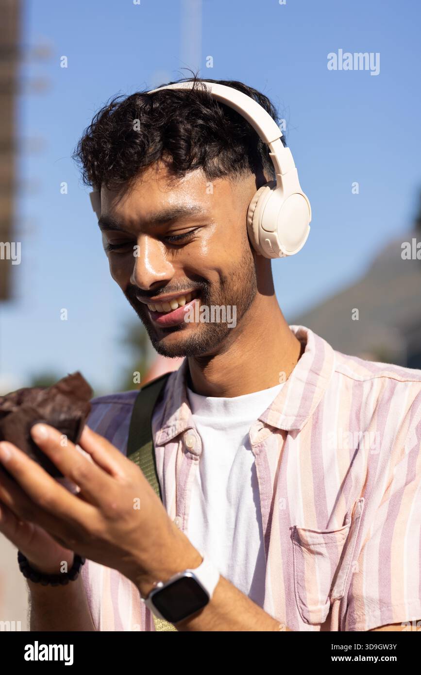 Homme asiatique souriant tout en tenant un muffin au chocolat, portant un casque circum-aural dans la rue de la ville Banque D'Images