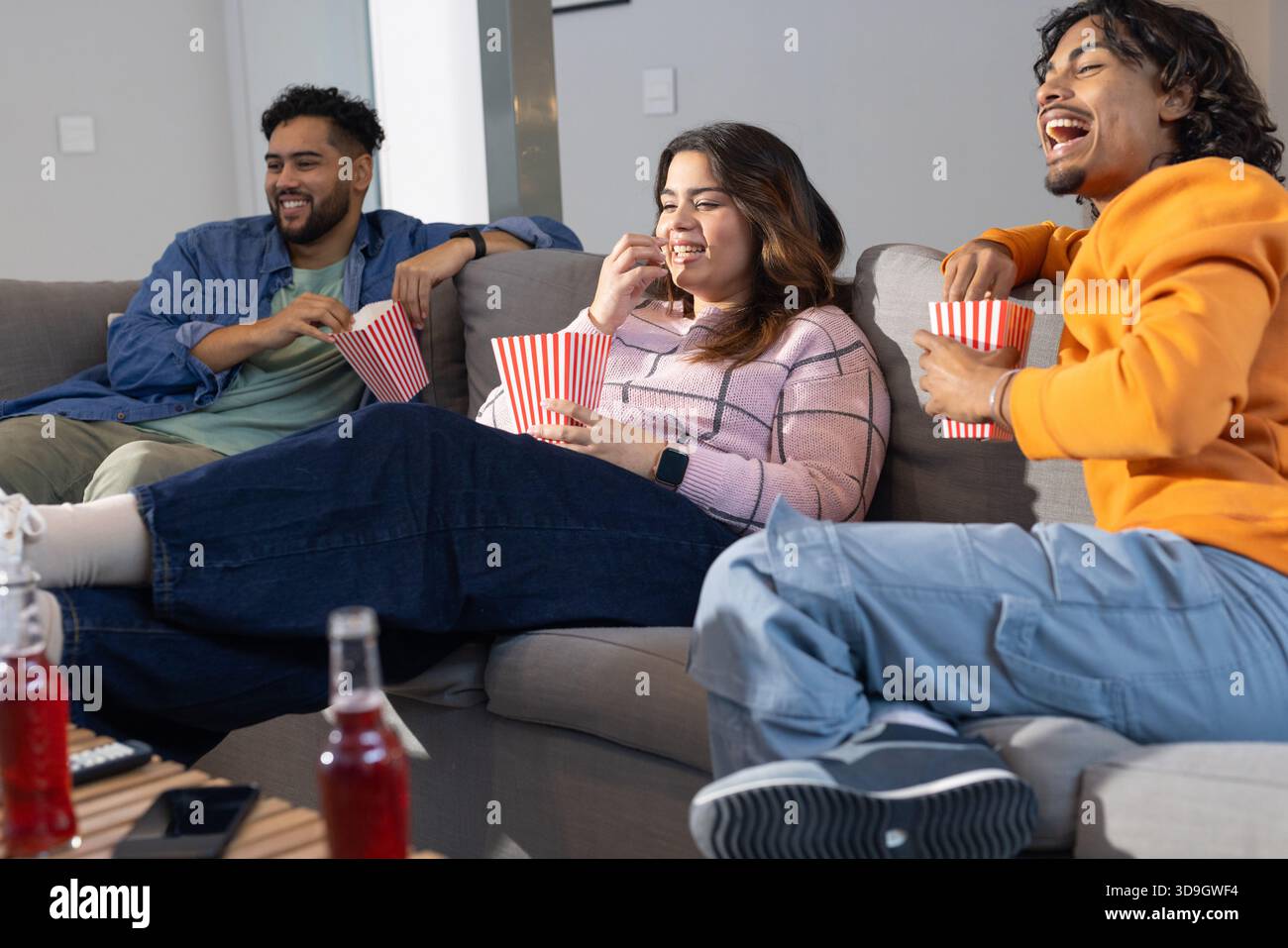 Divers amis regardant un film tout en étant assis sur un canapé gris clair dans le salon mangeant du pop-corn Banque D'Images