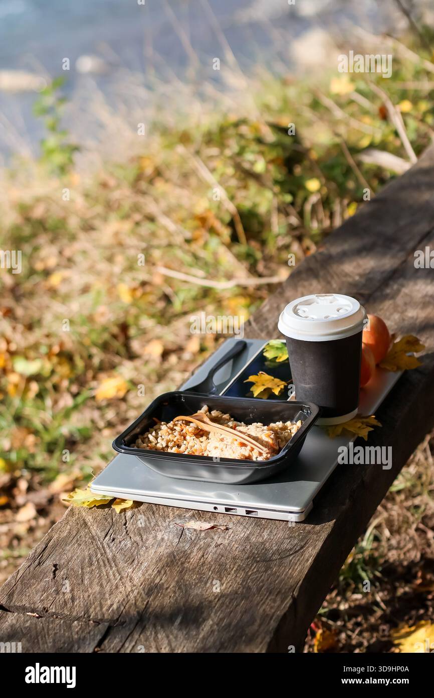 Plat de grain dans un récipient noir avec fourchette en bois, tasse à café à emporter, fruit orange, feuilles jaunes sur ordinateur portable argenté placé sur banc en bois donnant sur W. Banque D'Images