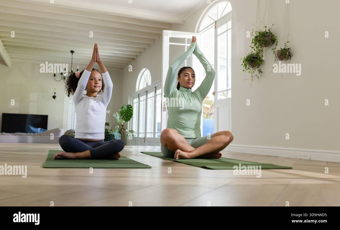 Mère et fille pratiquant le yoga levant les mains au-dessus de la tête sur des tapis dans le salon avec lustre Banque D'Images