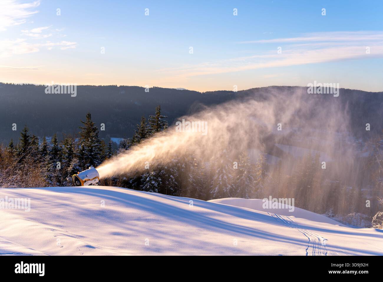 Un canon à neige fonctionne sur une pente enneigée dans une région montagneuse. L'appareil crée de la neige artificielle contre un ciel bleu clair. Les arbres bordent la scène, ajoutant au paysage hivernal. Banque D'Images