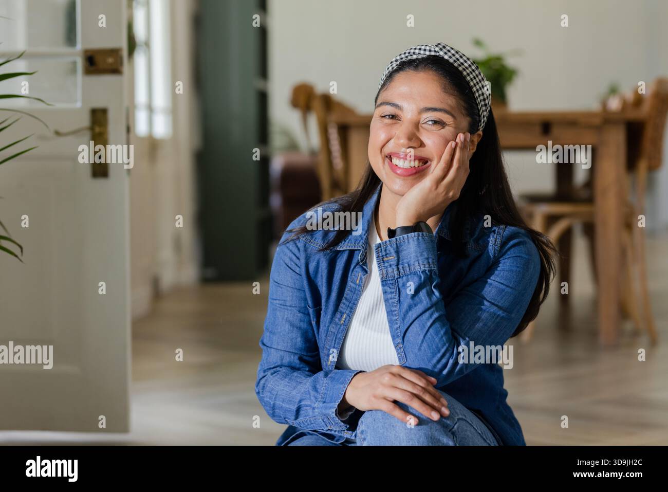 Femme se reposant joue sur la main tout en souriant et assise dans la salle à manger portant du denim et un bandeau Banque D'Images