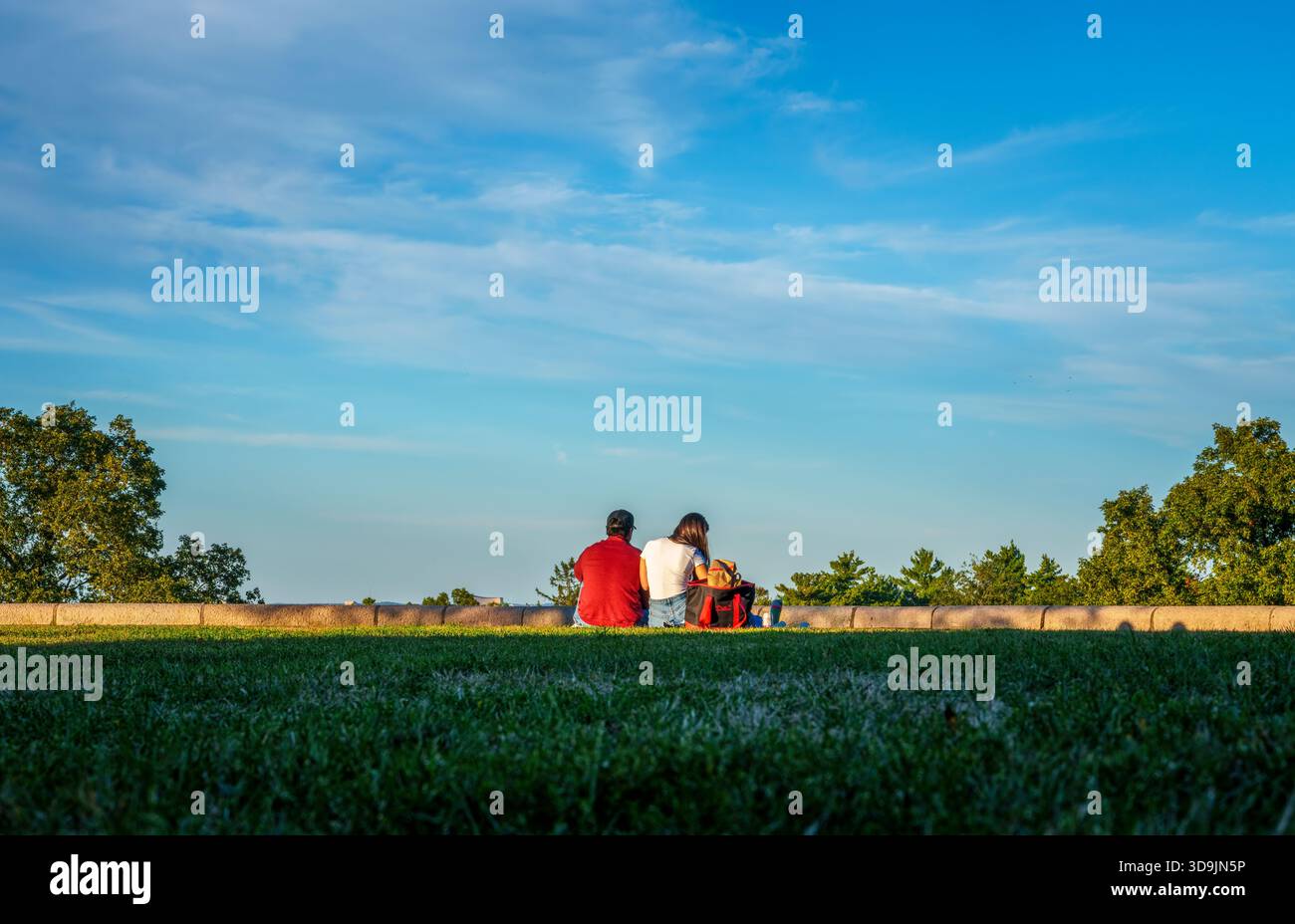 Jeune couple assis sur l'herbe avec un fourre-tout rouge et noir pique-nique au Larz Anderson Park à Brookline, ma, USA, sous un large ciel bleu du soir. Banque D'Images