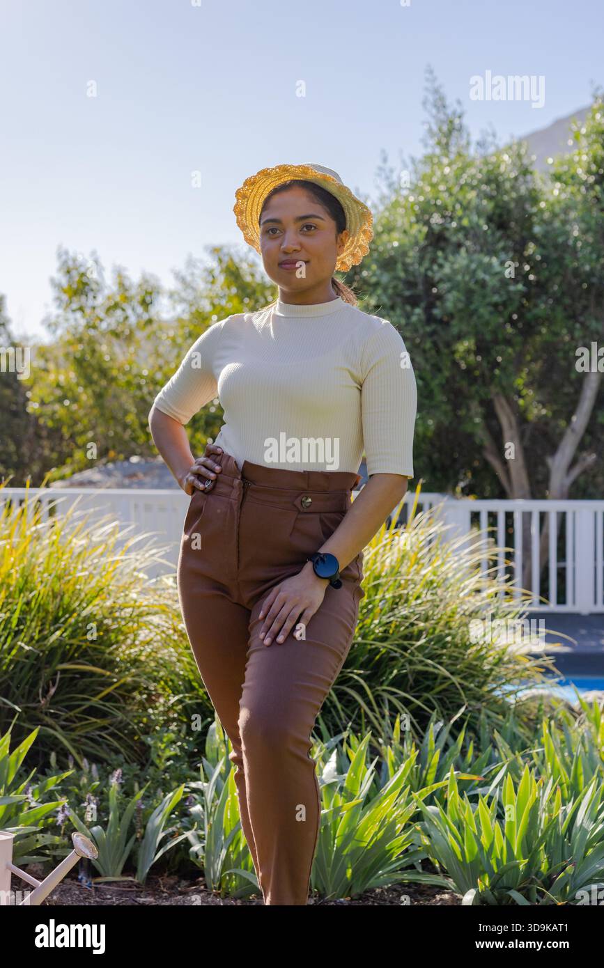 Femme debout dans la piscine arrière ensoleillée portant chapeau de paille et smartwatch près de l'herbe ornementale Banque D'Images