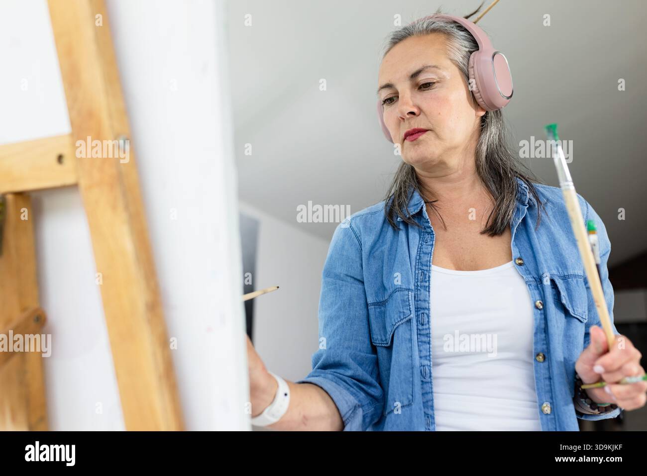 Peintre femme tenant des pinceaux peignant la toile tout en portant des écouteurs roses dans le studio d'art. Banque D'Images