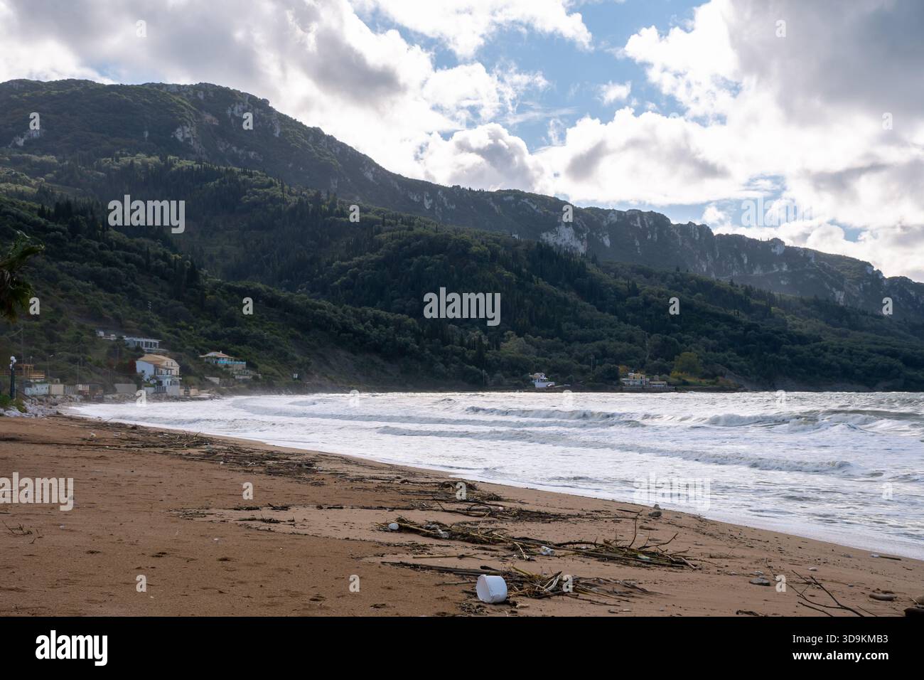 Vue grand angle d'une plage de sable couverte de débris de tempête et de bois flotté, avec des vagues rugueuses et une majestueuse montagne verte en toile de fond dans le nord de Corfou, GRE Banque D'Images