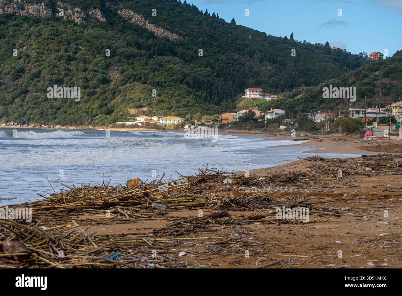 Vue grand angle d'une plage désordonnée couverte de milliers de bâtons de bambou et de litière en plastique après une tempête avec un village grec lointain niché dans le gr Banque D'Images