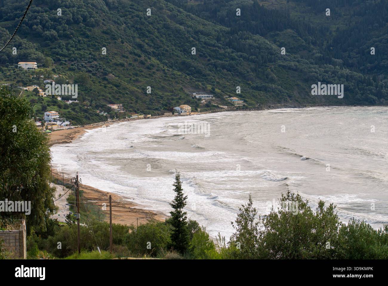Vue panoramique à angle élevé d'une grande baie en fer à cheval avec des vagues blanches rugueuses roulant sur le rivage et des montagnes verdoyantes parsemées de maisons sur un wi nuageux Banque D'Images