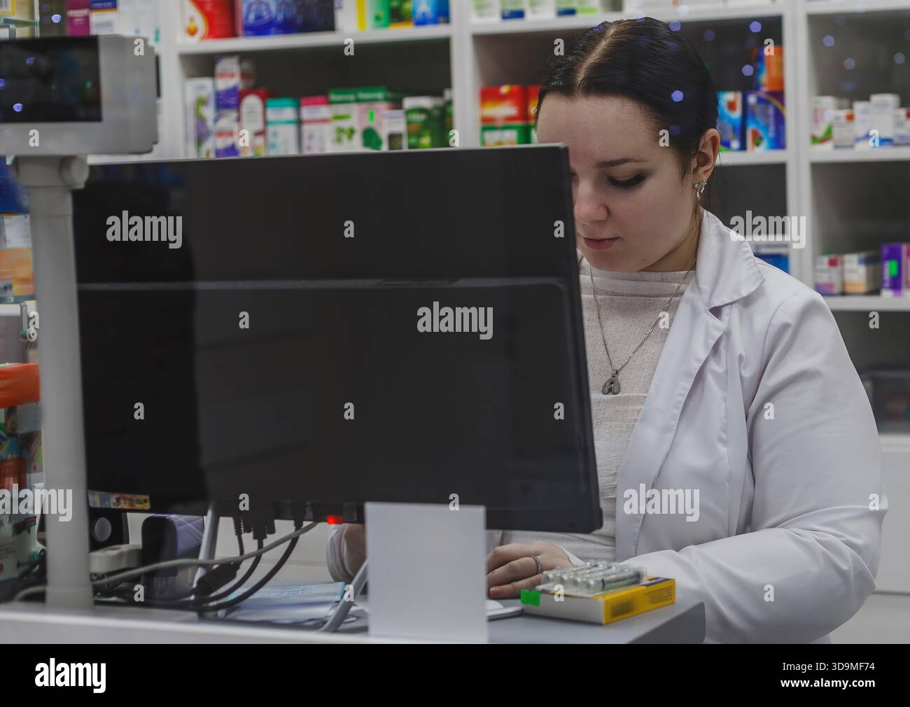 La photo montre une pharmacienne en blouse blanche traitant un achat de médicaments à un ordinateur, entourée d'étagères remplies de produits pharmaceutiques p Banque D'Images