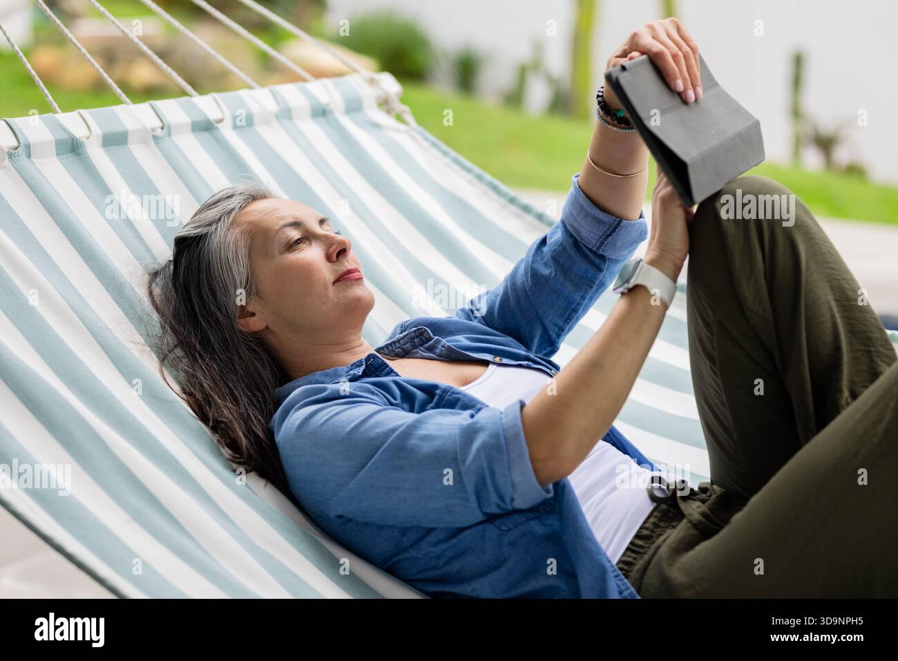 Femme adulte mature s'inclinant sur un hamac rayé dans la cour arrière tenant l'e-Reader noir au-dessus du visage Banque D'Images