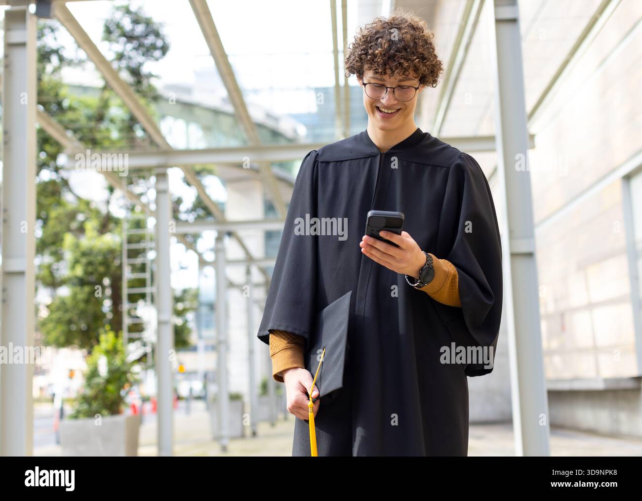 Diplômé masculin portant une robe debout sous la verrière sur le chemin du campus tenant smartphone et casquette Banque D'Images