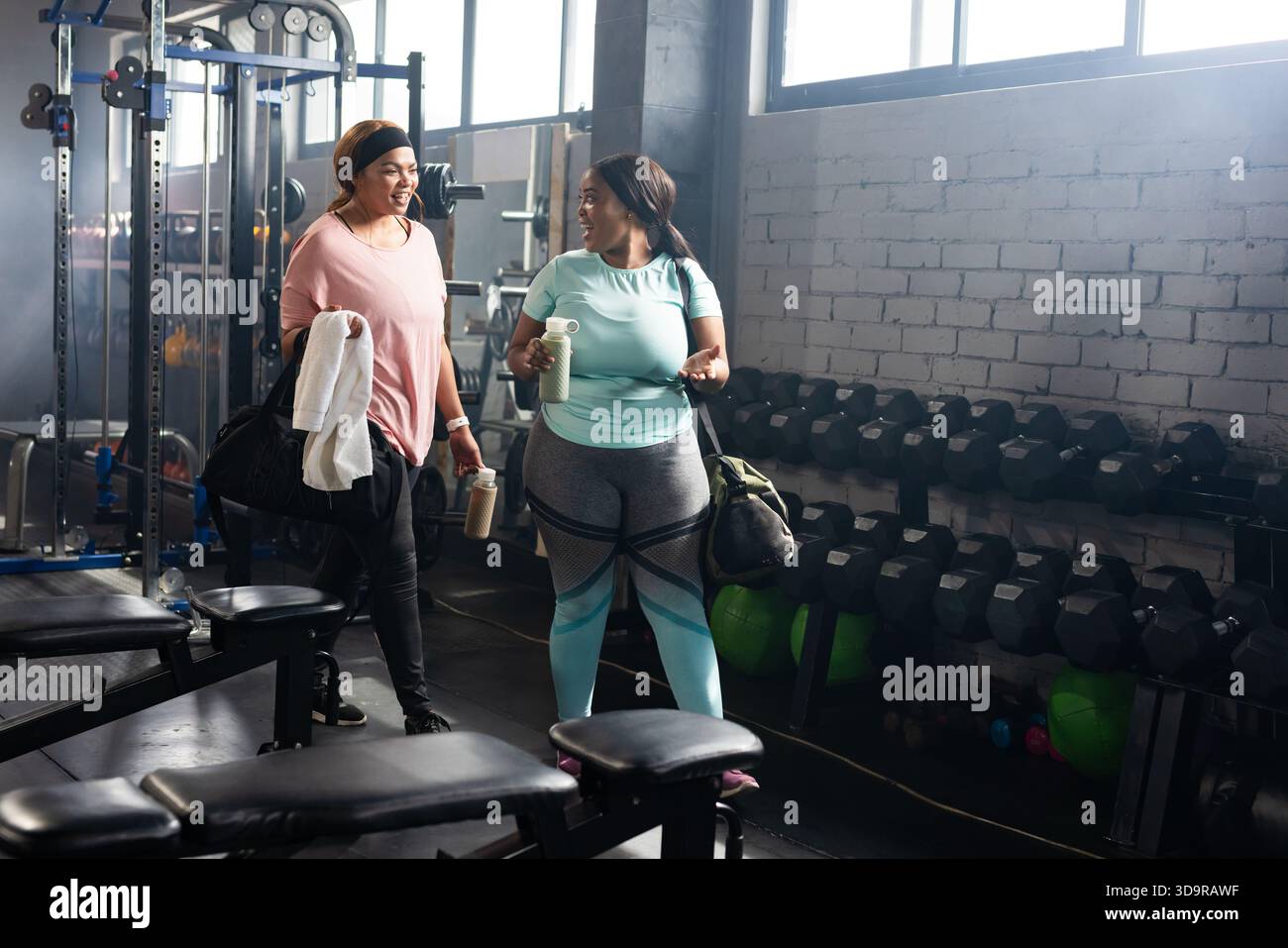 Diverses femmes adultes moyennes dans des vêtements d'entraînement tenant des bouteilles d'eau par porte-haltères dans le gymnase Banque D'Images