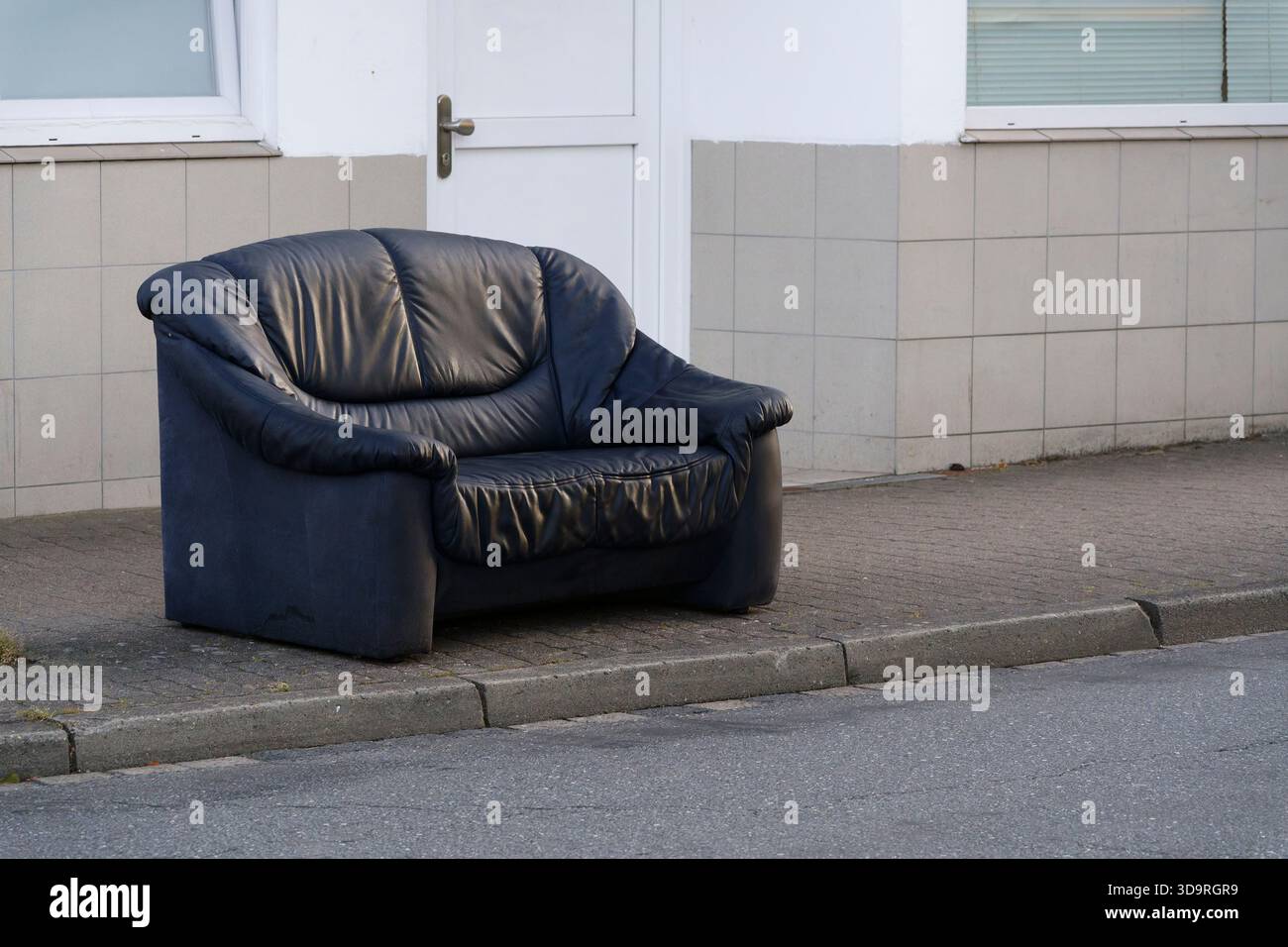 Sur le trottoir se dresse un vieux canapé deux places en cuir noir. Les meubles usés sont laissés à l'extérieur, montrant des signes d'âge et d'exposition aux intempéries dans une ville Banque D'Images