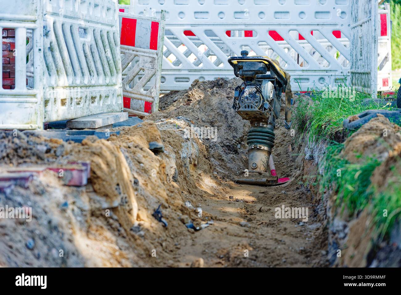 Une tranchée creusée se trouve derrière des barrières blanches avec des bandes rouges. Un compacteur de sol reste inactif à côté du gravier et des débris dispersés. L'herbe clairsemée borde la zone Banque D'Images