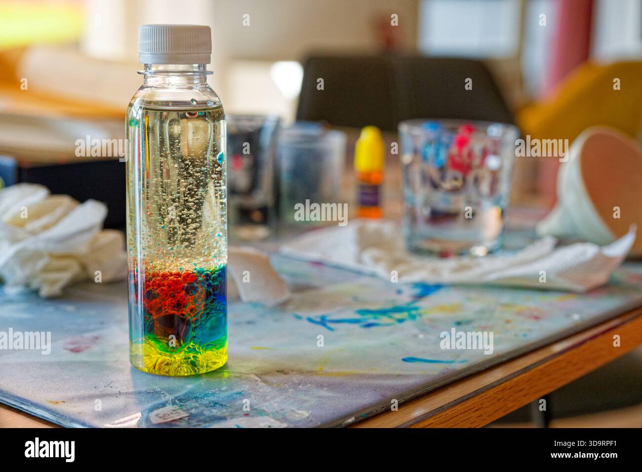 Une bouteille en plastique transparent contient des liquides empilés, jaune puis vert puis bleu puis rouge, une couche d'huile pâle flotte sur le dessus, des gouttes de peinture et des tissus reposent sur le Banque D'Images