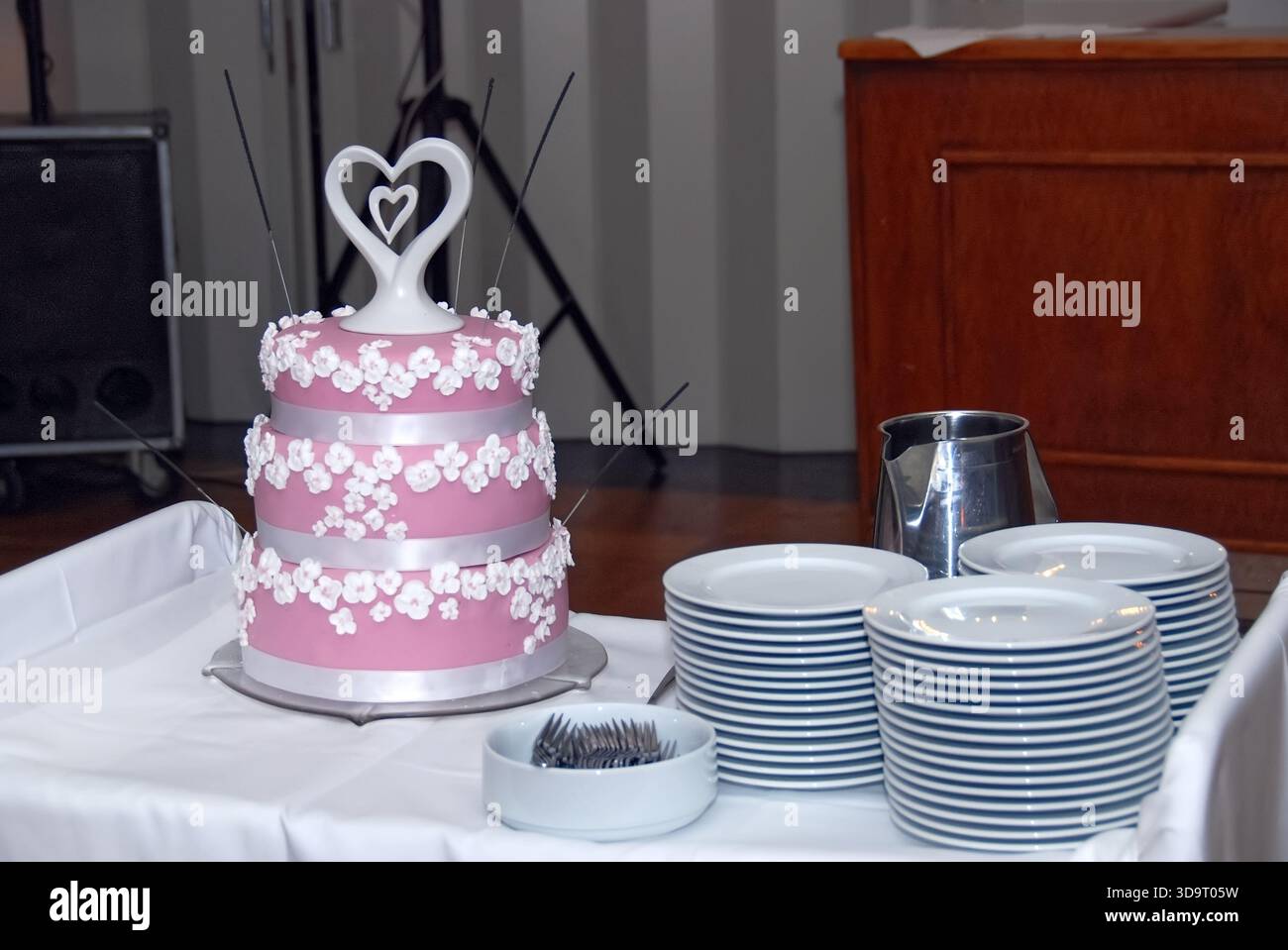 Un gâteau de mariage à trois niveaux magnifiquement décoré en rose avec des accents floraux blancs et un topper en forme de coeur, exposé sur une table avec des assiettes et uten Banque D'Images