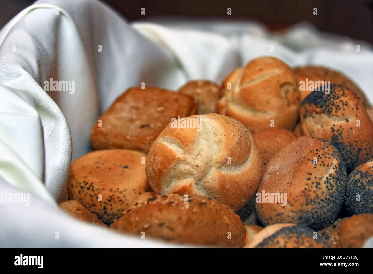 Un panier rempli de différents types de petits pains fraîchement cuits, y compris des variétés de graines de pavot et de graines de sésame, disposés soigneusement sur un chiffon blanc. Banque D'Images
