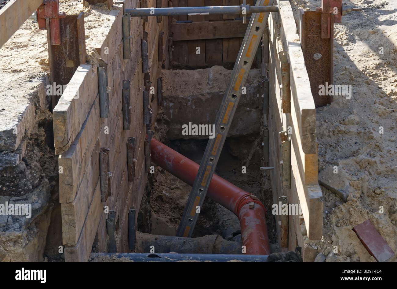 Une tranchée de construction avec des supports en bois et un tuyau rouge, avec une échelle pour l'accès à un chantier de construction à Hambourg aux beaux jours Banque D'Images