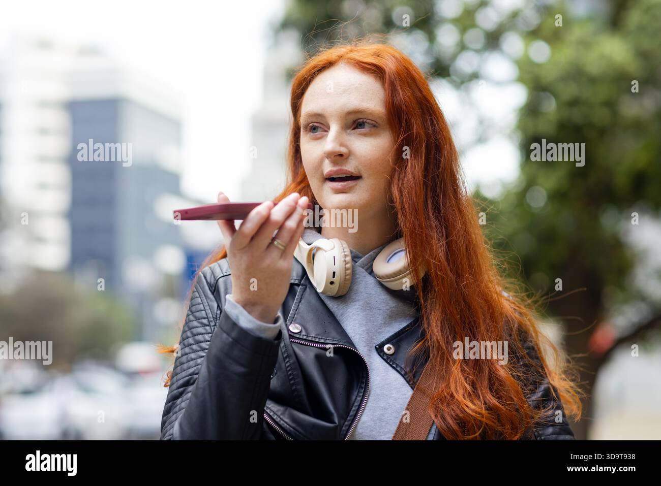 Femme aux cheveux rouges portant une veste en cuir et des écouteurs parlant dans un smartphone sur le trottoir de la ville Banque D'Images