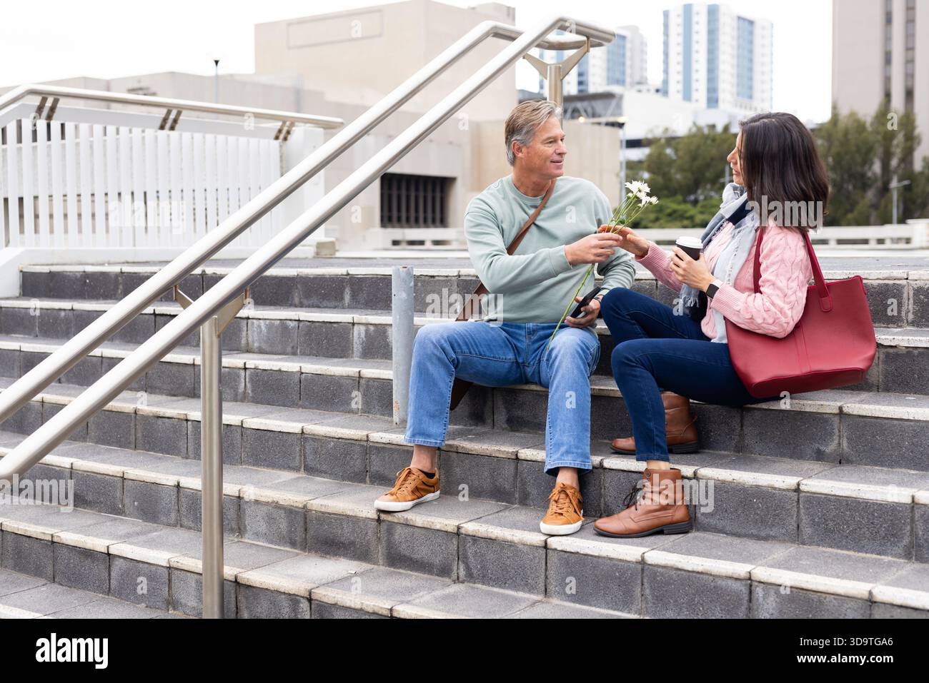 Couple diversifié assis sur des marches en béton dans la place urbaine échangeant marguerites blanches avec tasse à café Banque D'Images