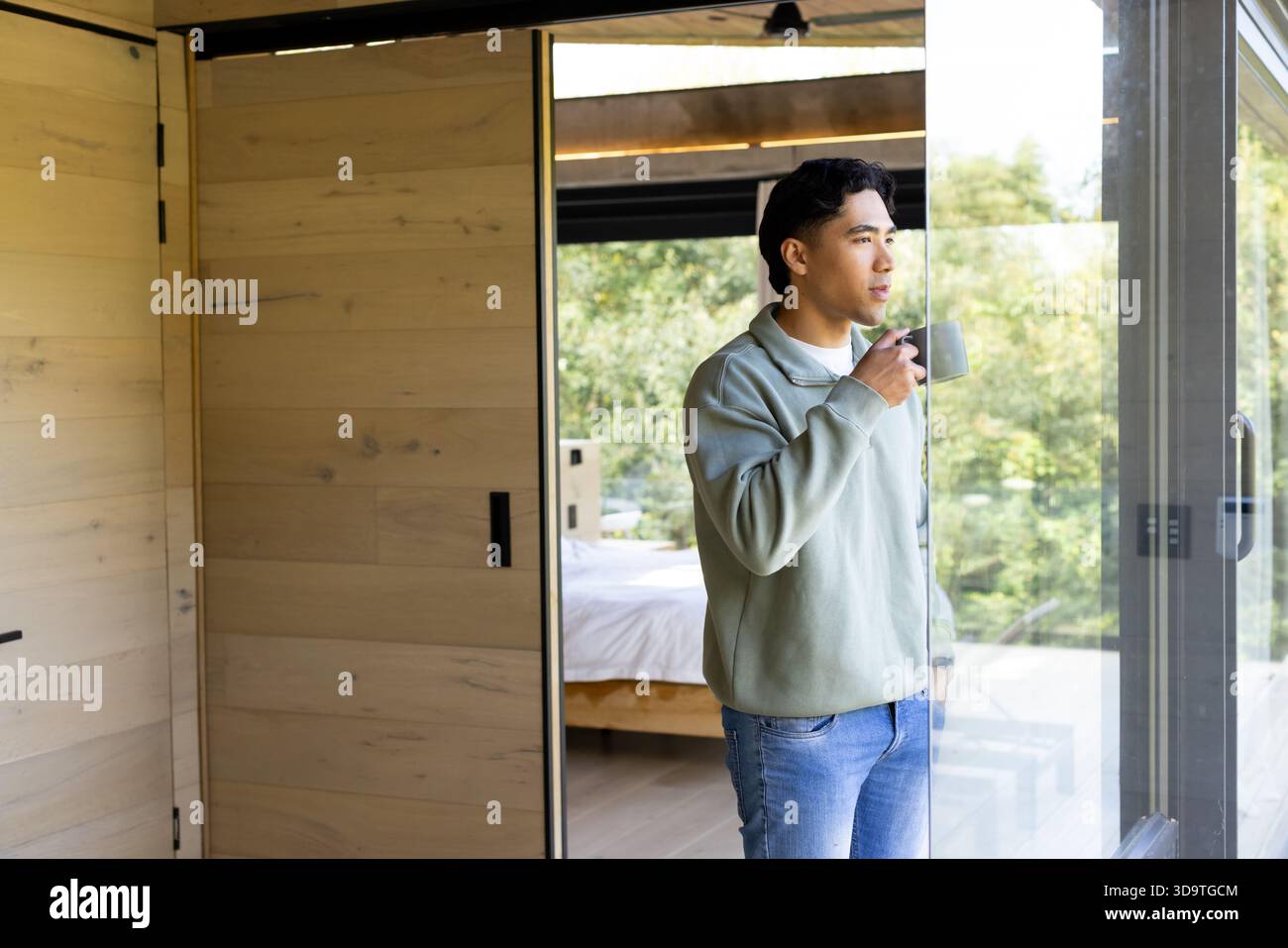 Homme asiatique debout dans la chambre tenant tasse à côté du lit regardant à travers la porte vitrée coulissante, espace de copie Banque D'Images