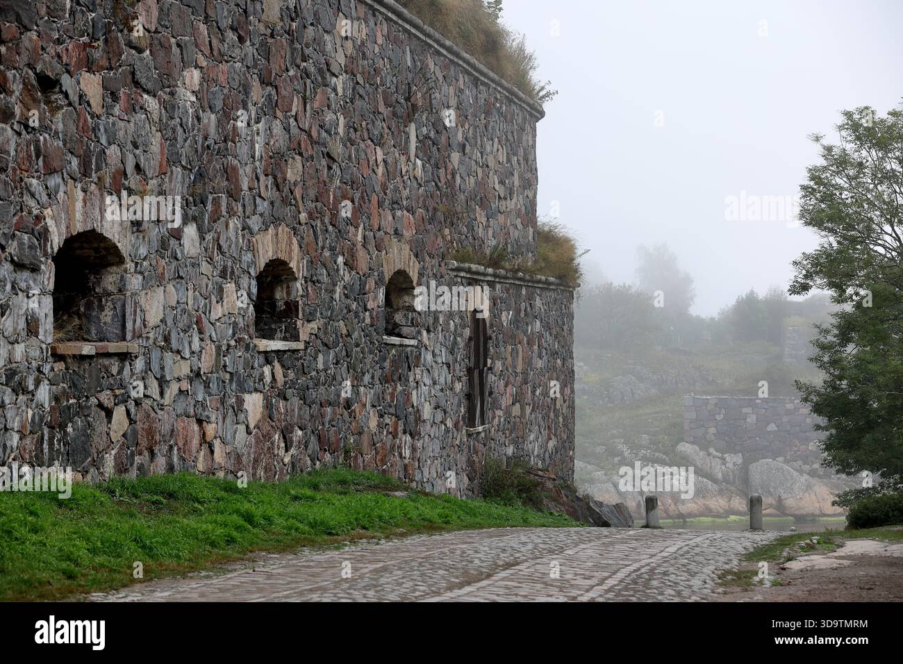 Embrasures arquées dans un mur de forteresse historique en pierre à côté d'un chemin pavé dans le brouillard par un matin brumeux à Suomenlinna Island, Helsinki, Finlande. Banque D'Images