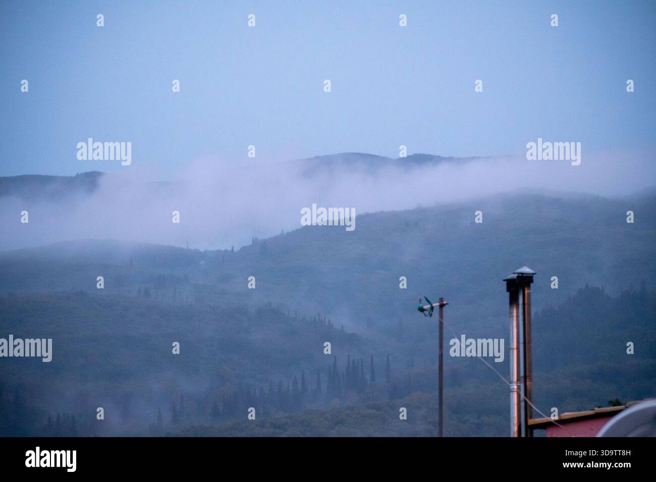 Paysage bleu atmosphérique avec brume roulant sur les collines et les montagnes forestières avec un tuyau de cheminée rural sur le toit et une antenne au premier plan Banque D'Images