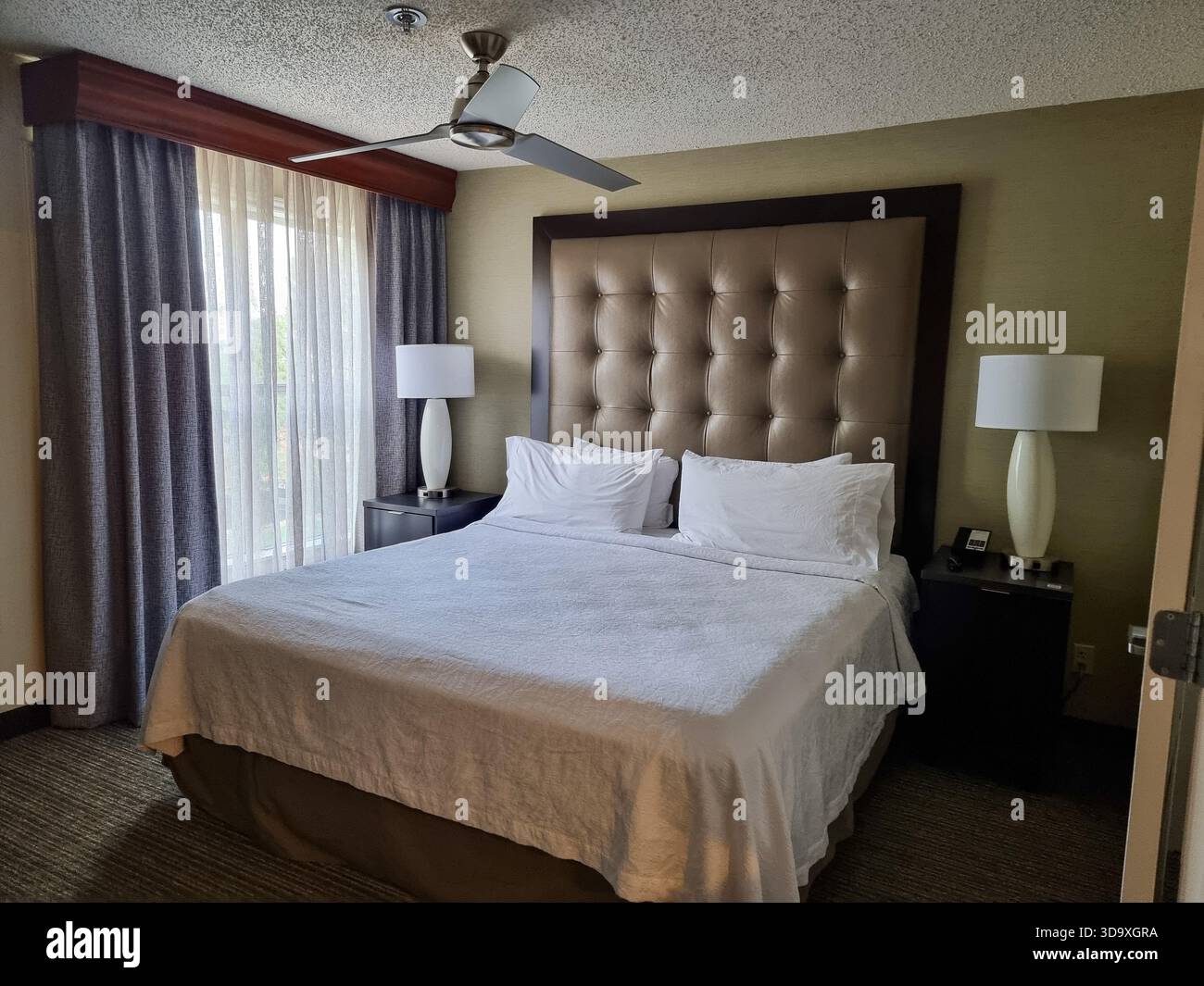 Chambre avec grand lit double / lit king près d'une fenêtre, ventilateur de plafond et détecteur de fumée dans un hôtel ; tête de lit en cuir brun, mur arrière rembourré - Image de stock capturée avec un smartphone