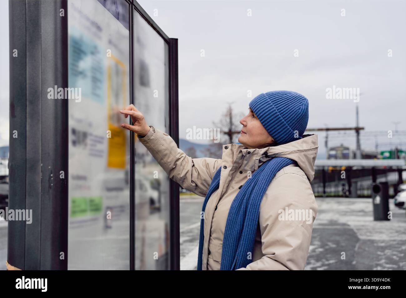 Adulte femme individu dans les vêtements d'hiver regarde le panneau d'information à l'extérieur, entouré de paysages urbains et ciel couvert, reflétant la communit Banque D'Images