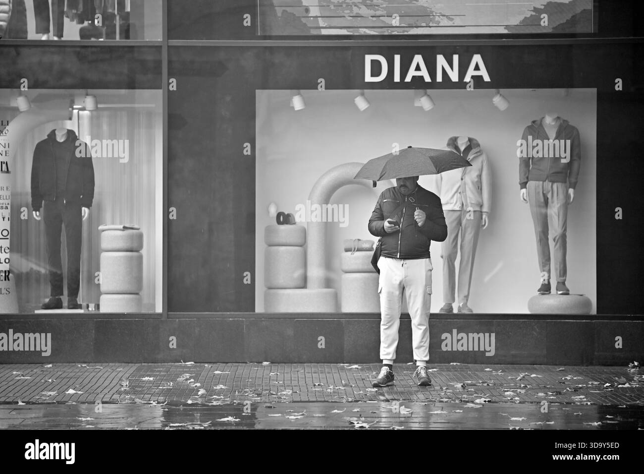 Erevan Arménie 12.7,2025. Un homme se tient debout sous la pluie avec un parapluie et un smartphone, passant par une fenêtre de magasin avec un panneau qui dit 'Diana' Banque D'Images