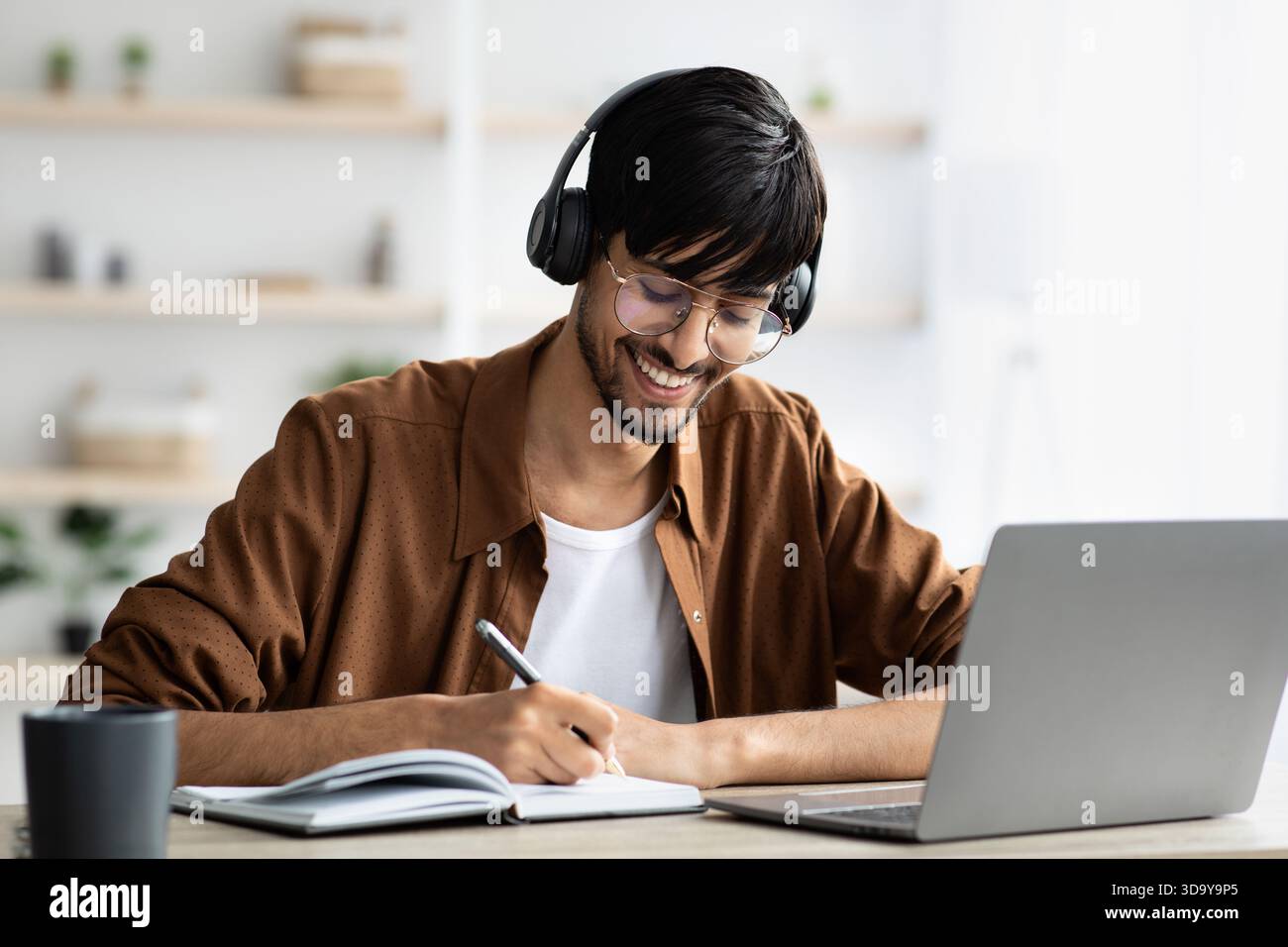 Homme étudiant à la maison avec des écouteurs tout en écrivant des notes dans un cahier et en utilisant un ordinateur portable dans une pièce bien éclairée Banque D'Images