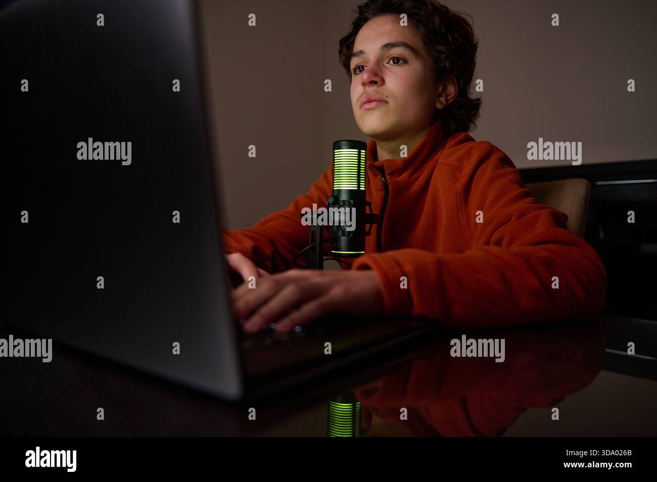 Un jeune dans une veste orange s'assoit à un bureau, tapant sur un ordinateur portable avec un microphone lumineux, créant une ambiance home studio calme pour le streaming ou la Rec Banque D'Images