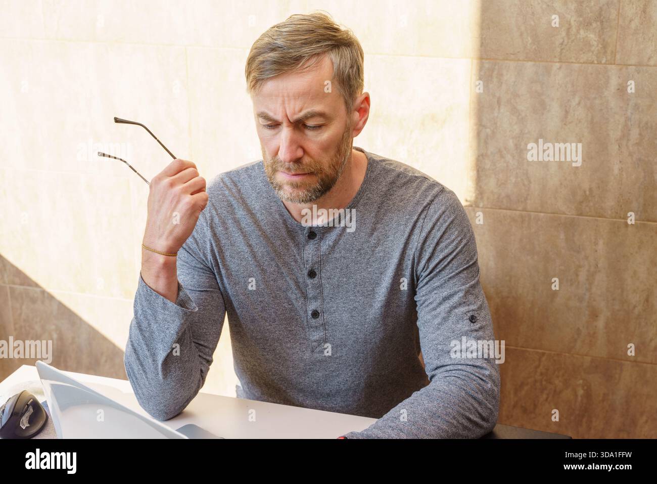 Un homme pensif d'âge moyen avec une barbe, portant une chemise grise, s'assoit à un bureau regardant vers le bas avec une expression concentrée tout en tenant ses lunettes. Banque D'Images